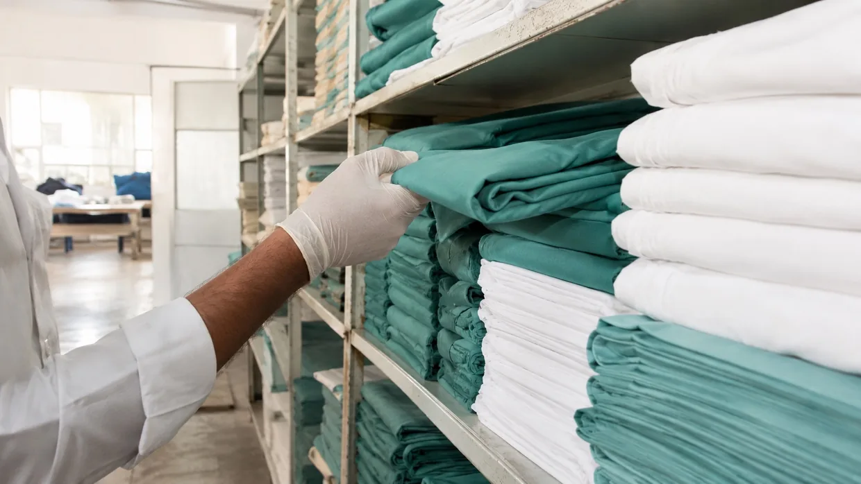 A gloved hand pulling a bolt of teal scrub fabric from a shelf of folded medical fabric