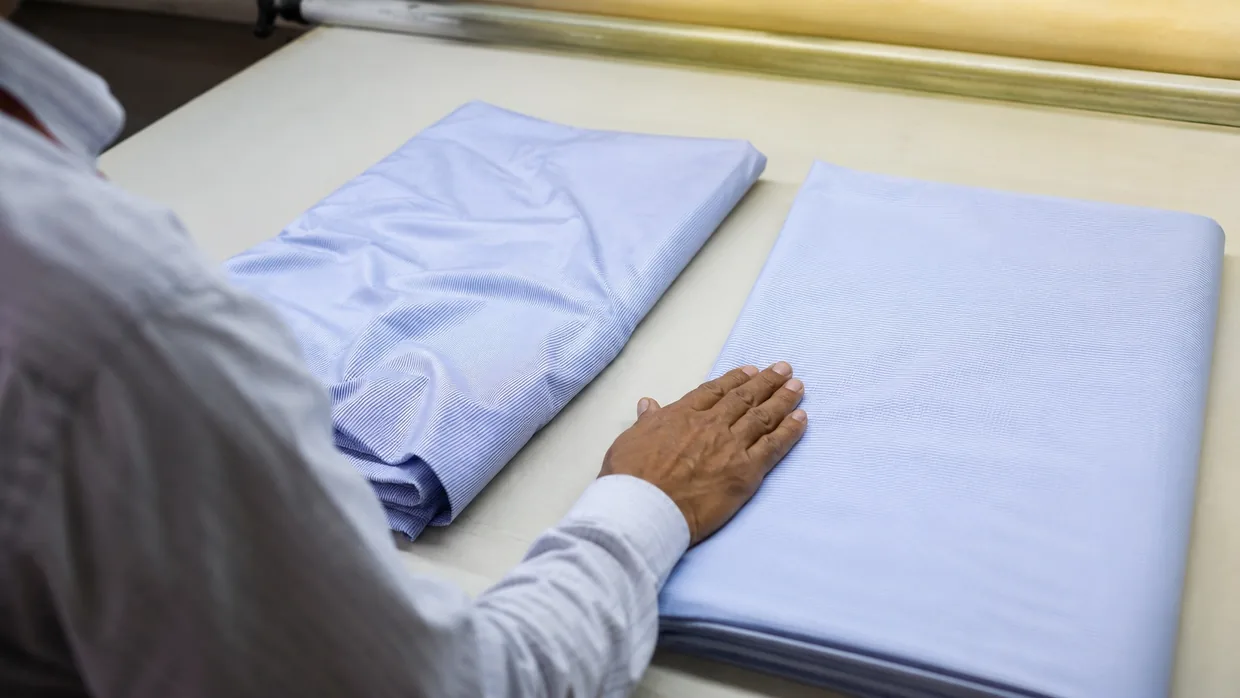 Two pale shirting swatches compared side by side, one creased and one smooth, on a mill table