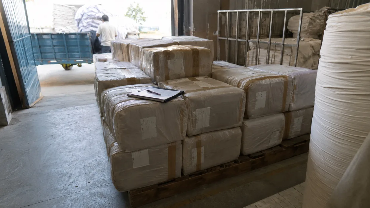 Taped and labelled fabric parcels stacked on a pallet in a dispatch bay with a clipboard on top